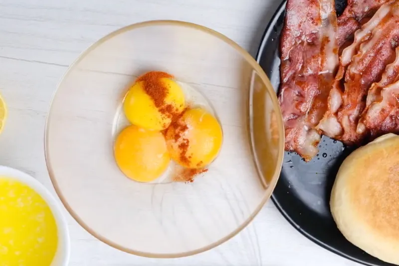 Three raw egg yolks in a glass bowl seasoned with paprika and salt, next to melted butter, lemon, crispy bacon and English muffins