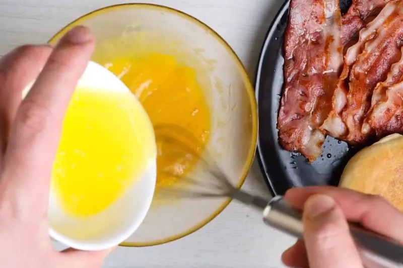 Melted butter being poured into whisked egg yolks in a glass bowl to make hollandaise sauce, with crispy bacon and an English muffin nearby