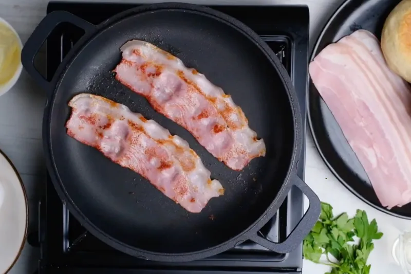 Two strips of bacon frying in a black skillet, with raw bacon, an English muffin and fresh parsley on the counter beside it