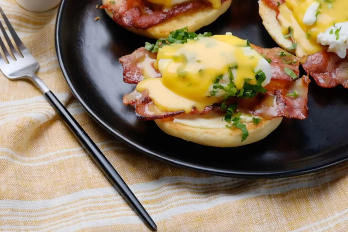 Siyah tabakta servis edilen, kızarmış İngiliz kekinin üzerinde çıtır bacon, hollandaise sos ve doğranmış maydanozla hazırlanmış Benedict yumurta